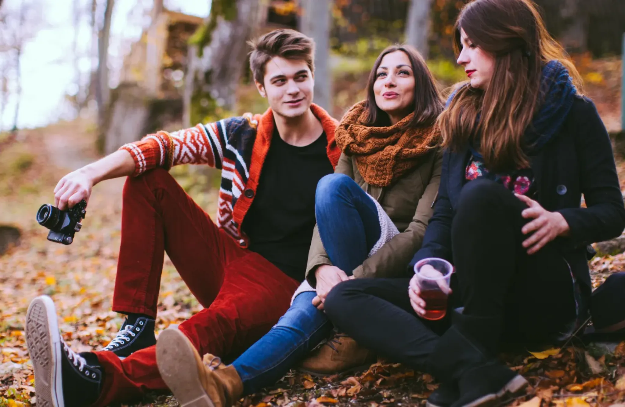 Three people sitting on the ground outdoors in autumn, two women and one man, talking and holding a drink.